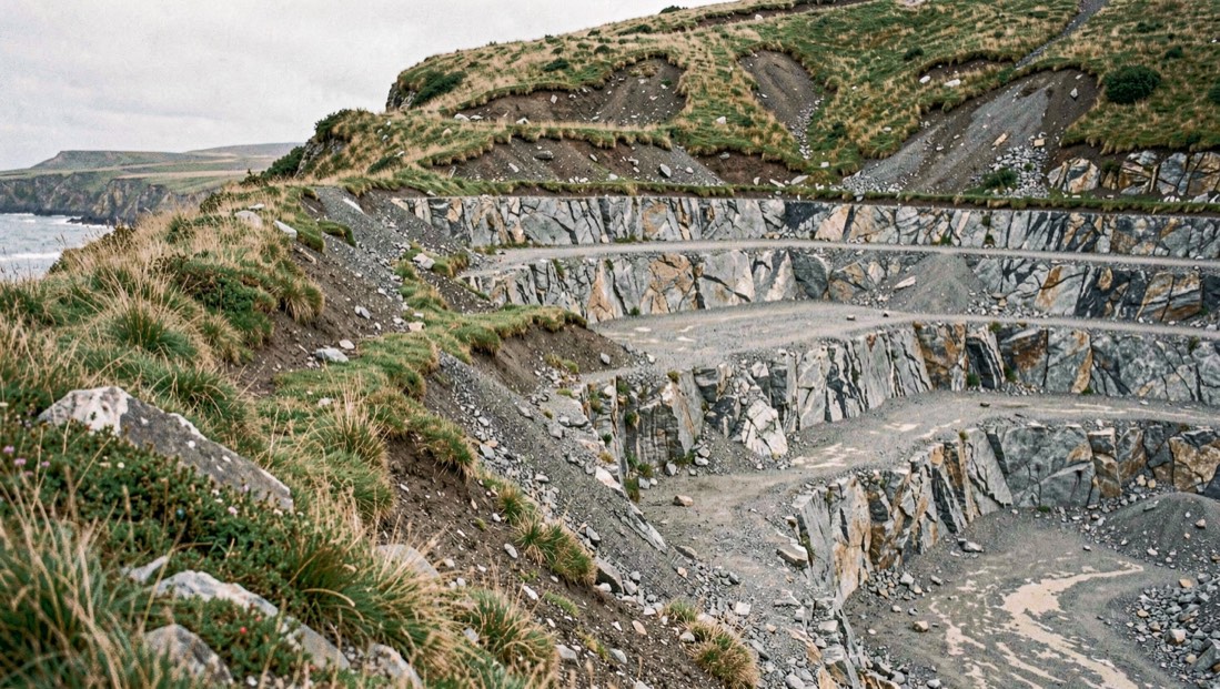 Stepped spoil terraces of a former alum quarry cut into a coastal hillside with rough grass and exposed grey shale